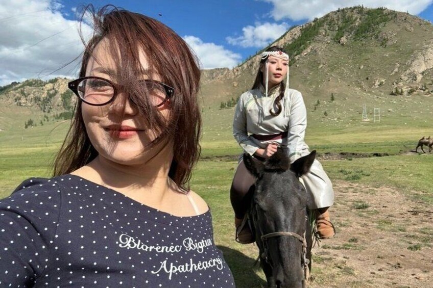 A person in a polka dot shirt and another in traditional attire on horseback in a grassy field with a mountain in the background. 3