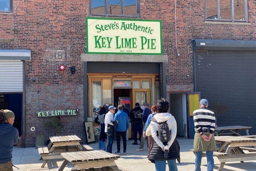 A brick building with a green sign for 'Steve's Authentic Key Lime Pie.' People are standing outside, and there are wooden tables in front. 3