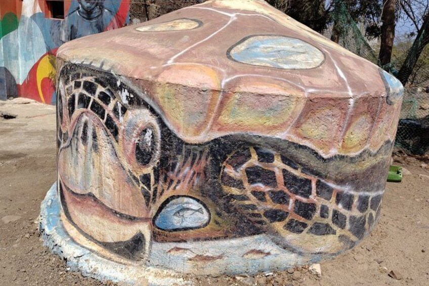 A large turtle sculpture with a mosaic pattern, surrounded by a sandy area and colorful murals in the background. 2