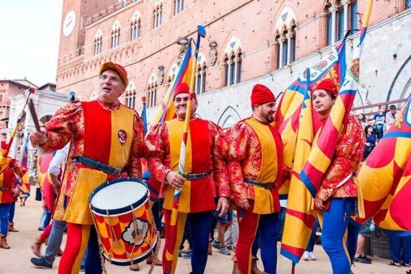A group of people in medieval costumes with flags, a building with arched windows, and a clock tower in the background. 4