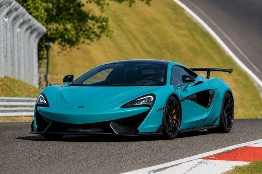 A teal sports car on a race track with a grassy hill and a guardrail in the background. 3