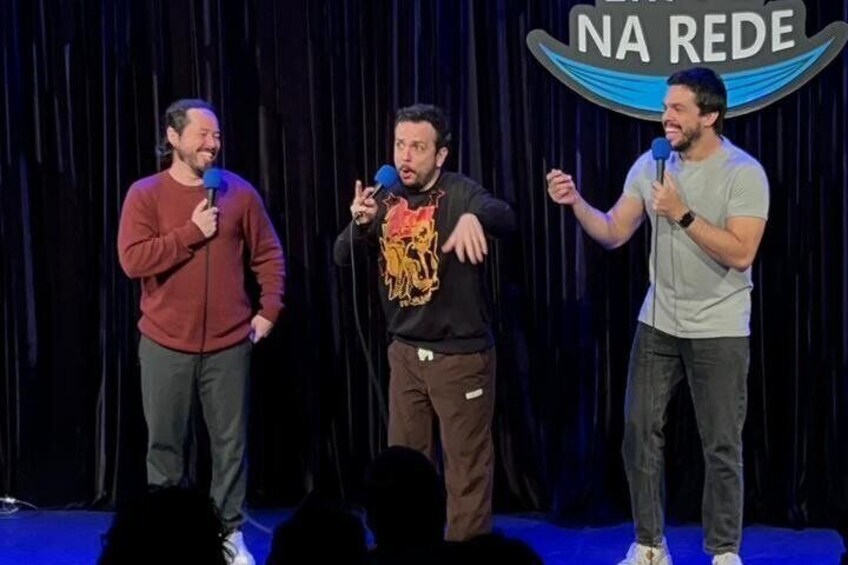 Three men on stage with microphones, a sign that reads 'NA REDE' in the background. 1