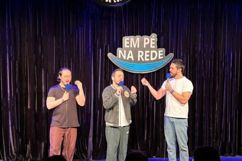 Three people on stage with microphones, a sign that reads 'EM PE NA REDE', and a dark curtain background. 3