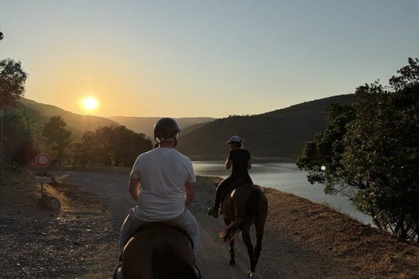 Two people horseback riding along a dirt path beside a body of water during sunset. 4