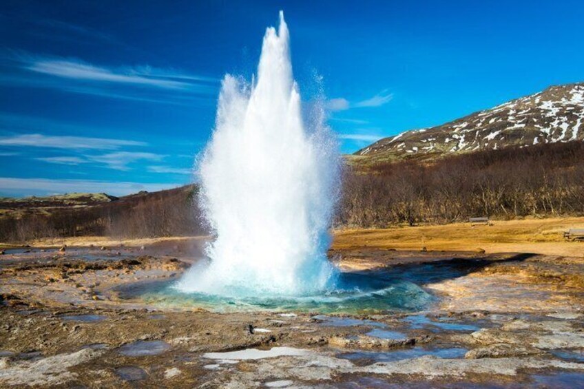 A geyser erupting in a natural landscape with a mountain in the background. 1