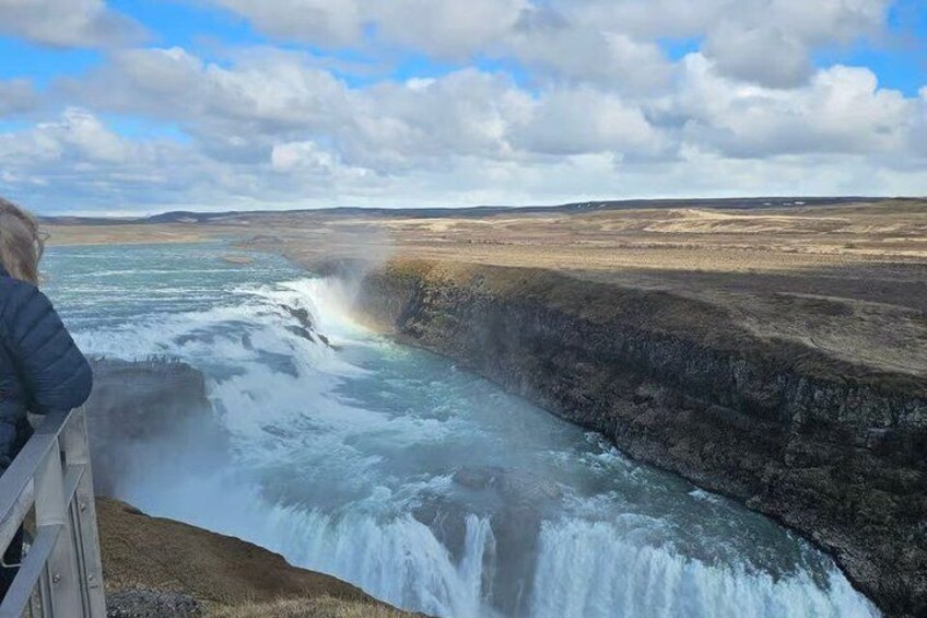 A waterfall cascading over a rocky cliff, a person observing from a wooden railing, and a vast, open landscape under a partly cloudy sky. 3