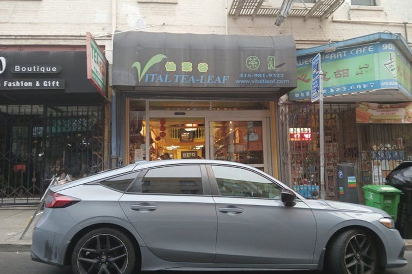 A grey car parked in front of a shop named 'ITAL Tea-Leaf' with a signboard and a storefront. 3