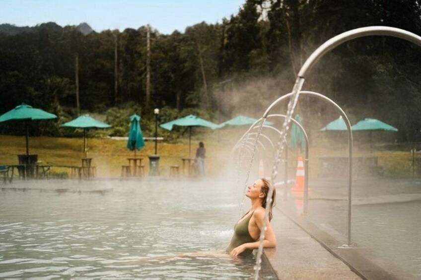 A person relaxing in a hot spring with steam rising, surrounded by green umbrellas and chairs. 4