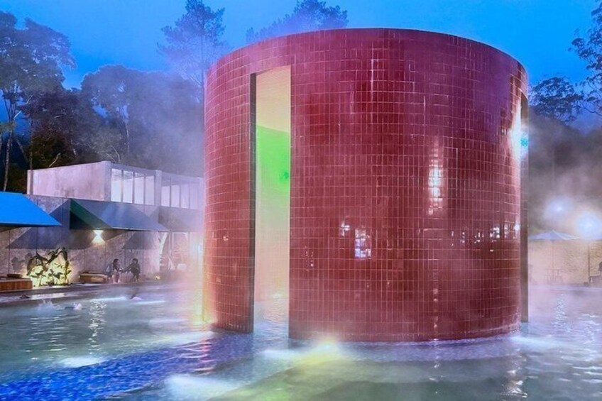 A cylindrical structure with a reflective surface, surrounded by water and mist, with a geometric building in the background. 3