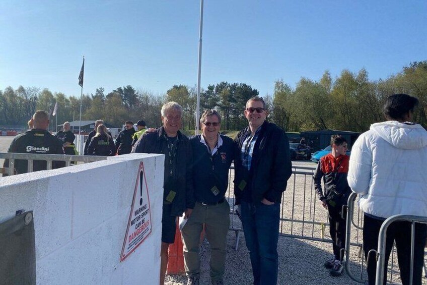 A group of people standing near a white wall with a warning sign, a flag, and a metal fence in the background. 3