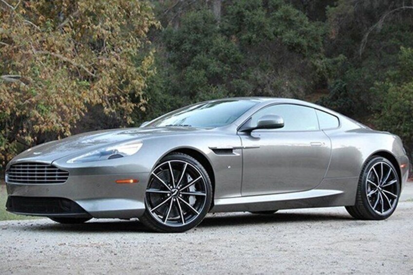 A silver sports car parked on a gravel surface with trees in the background. 1