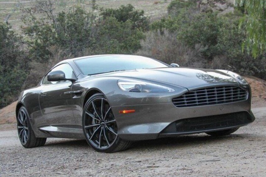 A silver sports car parked on a gravel surface with a natural landscape in the background. 3