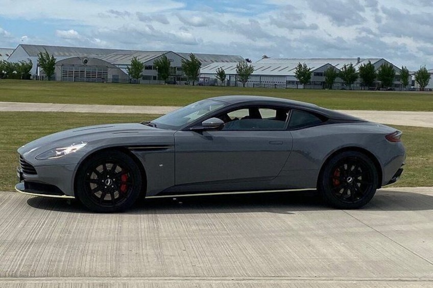 A gray sports car parked on a concrete surface with a grassy field and industrial buildings in the background. 5