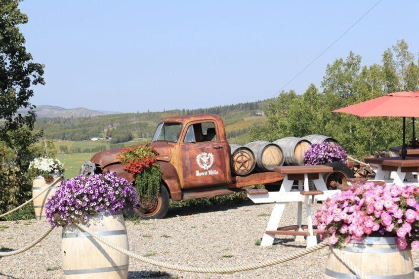 An old rusty truck with barrels and flowers, set in a rural landscape with trees and hills. 1