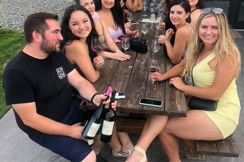 A group of people sitting at a wooden table outdoors, holding wine glasses and bottles. 2