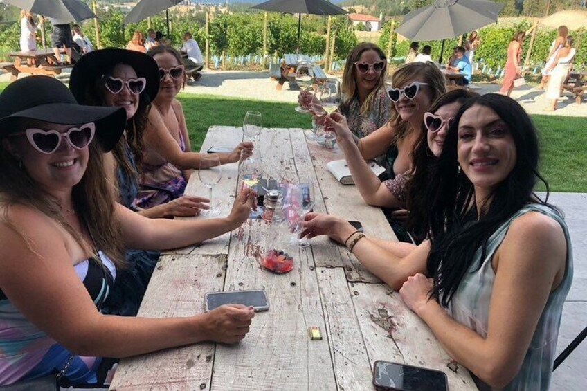A group of people sitting at a wooden table outdoors, holding drinks and toasting. 3