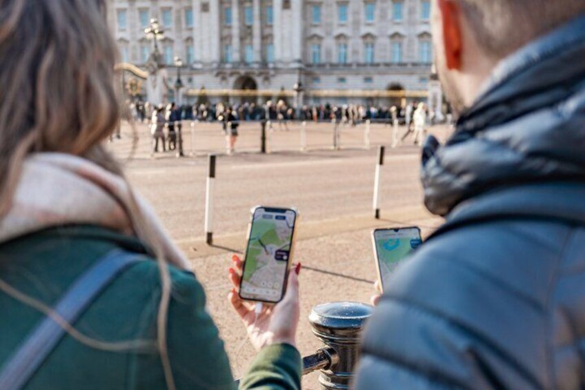 Two people using smartphones, possibly for navigation, in a public square with a historic building in the background. 2