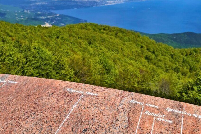 A granite monument with inscriptions, a forested hillside, and a distant body of water. 4