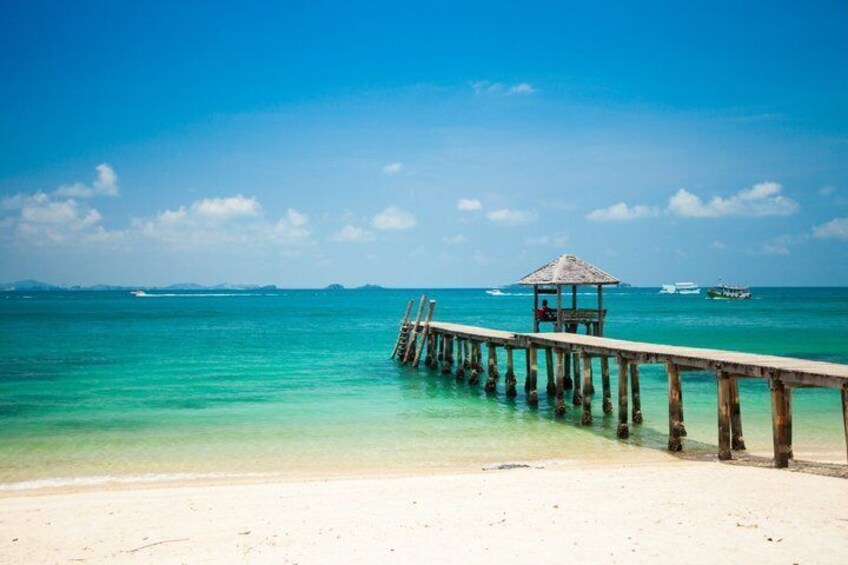 A beach with a wooden pier extending into turquoise water, a thatched-roof structure at the end, and boats in the distance. 3