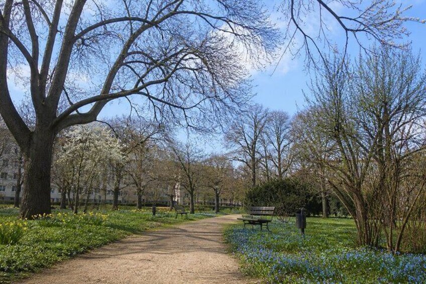 A park with a dirt path, trees, a bench, and blooming flowers. 2