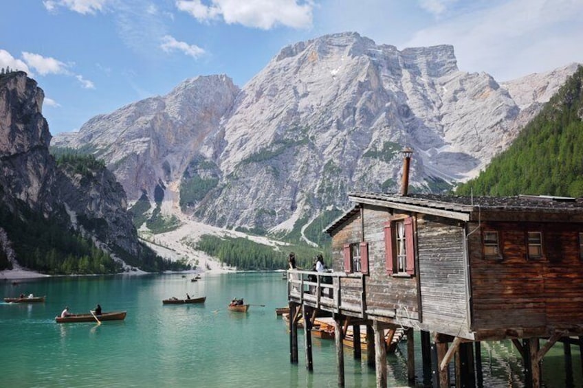 A wooden house on stilts by a turquoise lake with boats, surrounded by mountains. 1