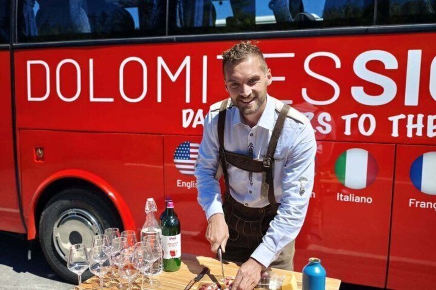 A man serving drinks at a table with wine glasses and bottles in front of a red bus with language options displayed. 4