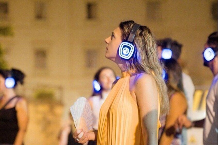 A person in a yellow dress holding a white object, with headphones on, in a crowded outdoor setting. 1
