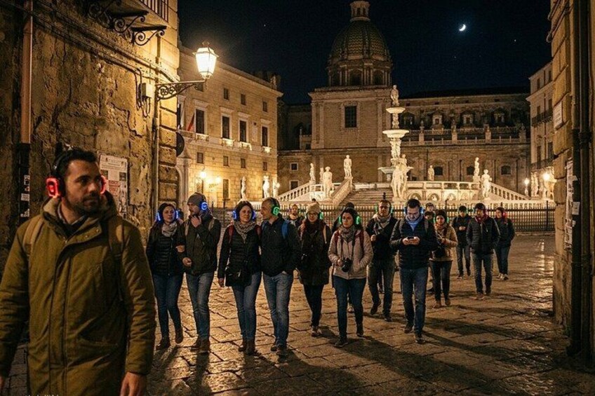 A group of people walking on a cobblestone street at night with illuminated buildings and a fountain in the background. 2