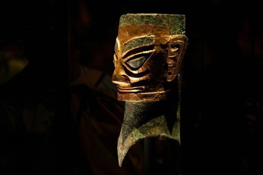 A carved wooden mask with a prominent eye and nose, set against a dark background. 2