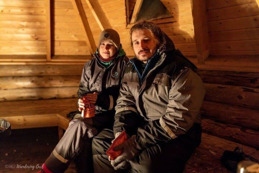 Two people sitting in a wooden cabin with a log wall and a bench. 4