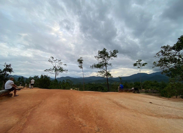 A dirt road with people sitting and standing, surrounded by trees and mountains under a cloudy sky. 3