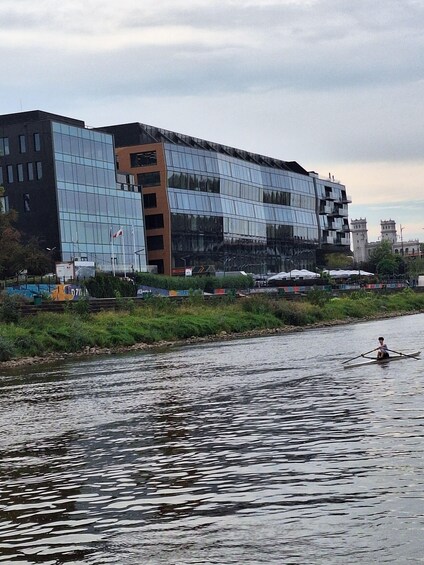 A modern building with reflective glass windows, a river with a person rowing, and a cloudy sky. 3