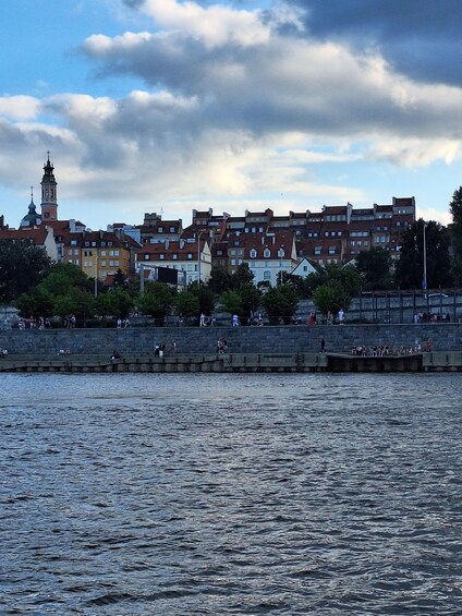 A waterfront cityscape with historic buildings, a prominent tower, and a stone embankment with people. 4