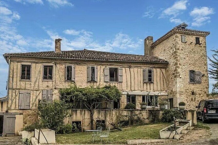 A large stone house with a tower, surrounded by a well-maintained garden and a parked car. 1