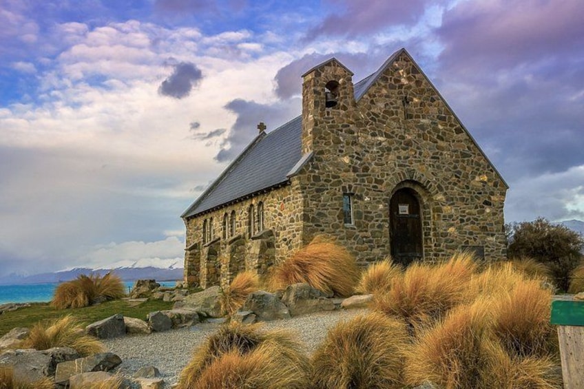 A stone church with a steep roof, surrounded by grassy terrain and a body of water in the background. 5