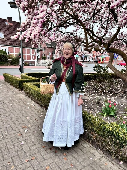 A person in traditional attire standing in a park with blooming trees and a brick building in the background. 3