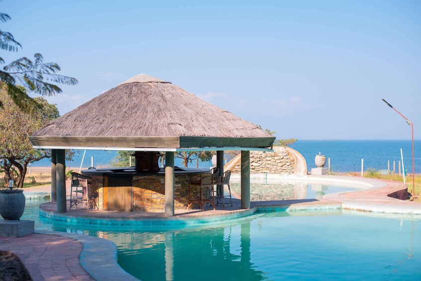 A pool with a thatched gazebo, stone pillars, and a view of the sea. 3