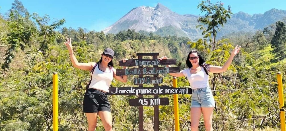Two people standing in front of a signpost with text and a mountain in the background. 2