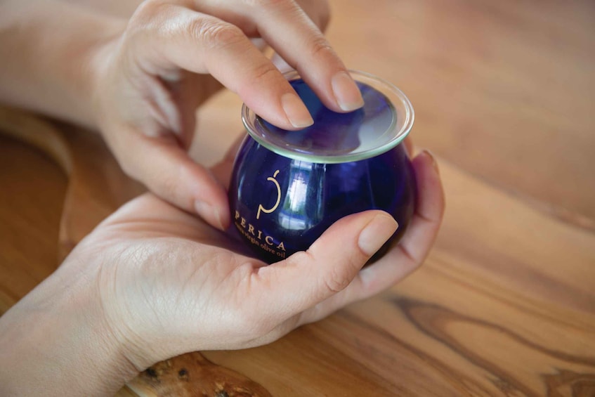 A person holding a small blue container with the label 'PERICA' and 'extra virgin olive oil' on a wooden surface. 5