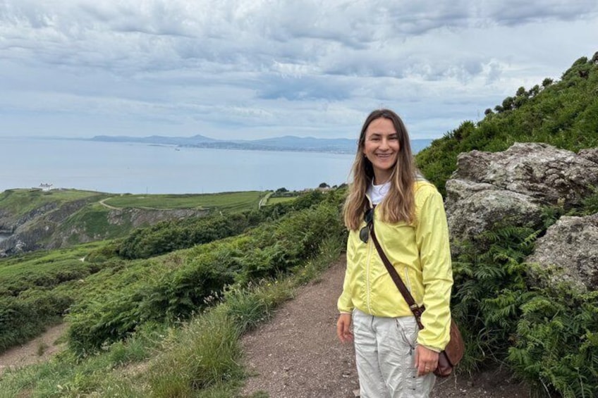 A person standing on a grassy path with a view of the sea and hills in the background. 4