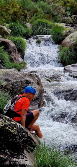 A person in a red shirt and blue helmet crouching on a rock by a flowing river. 1