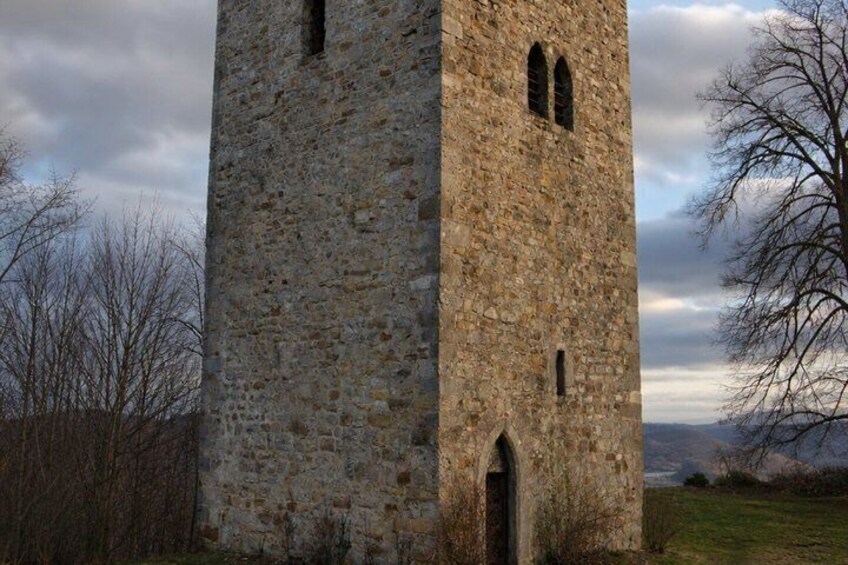 A tall stone tower with arched windows and a door, surrounded by a grassy landscape and bare trees under a cloudy sky. 2