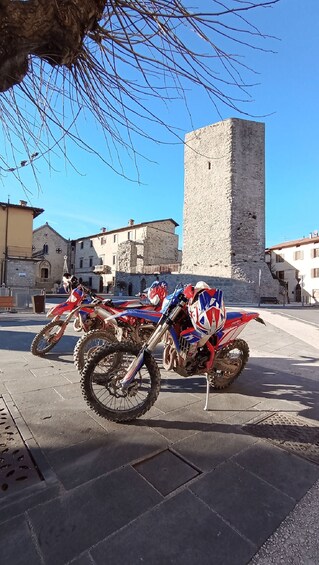 A row of dirt bikes parked on a paved area with a stone tower in the background. 4