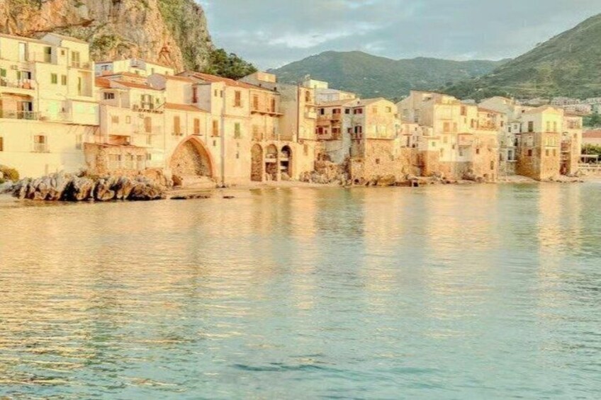 A coastal town with buildings along the water, a rocky cliff, and mountains in the background. 2