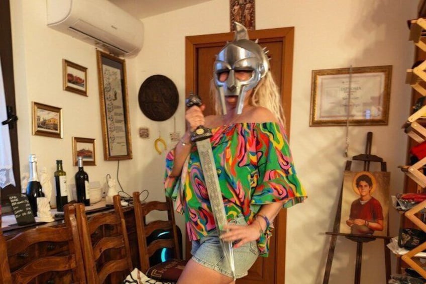 A person in a colorful off-shoulder top and shorts, holding a sword, standing in a room with wooden furniture, a painting, and various wall decorations. 4