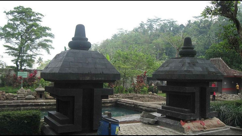 Two stone structures with pointed tops, a small pond, and lush greenery in the background. 2