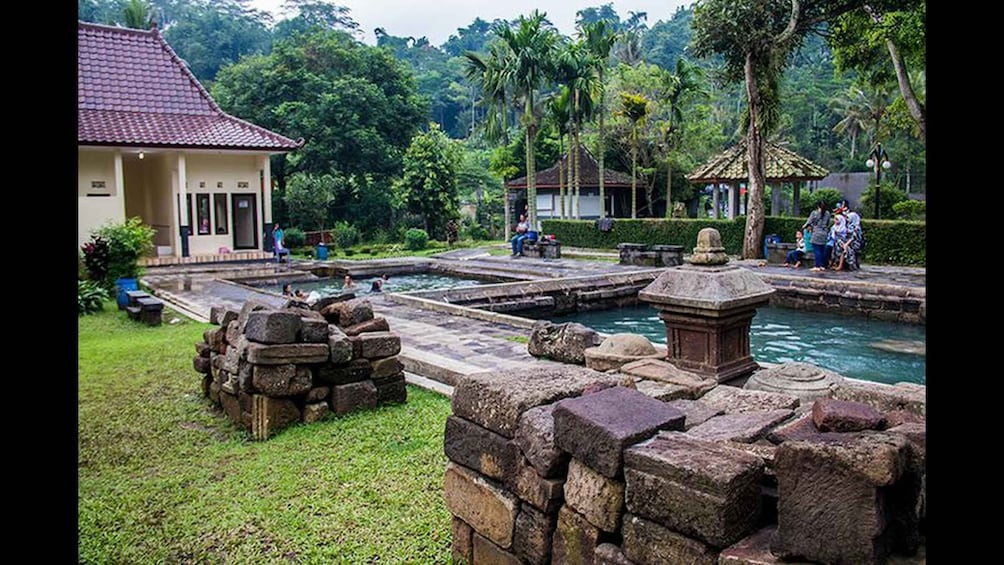 A traditional stone structure with a tiled roof, a stone wall, and a water feature with people around. 4