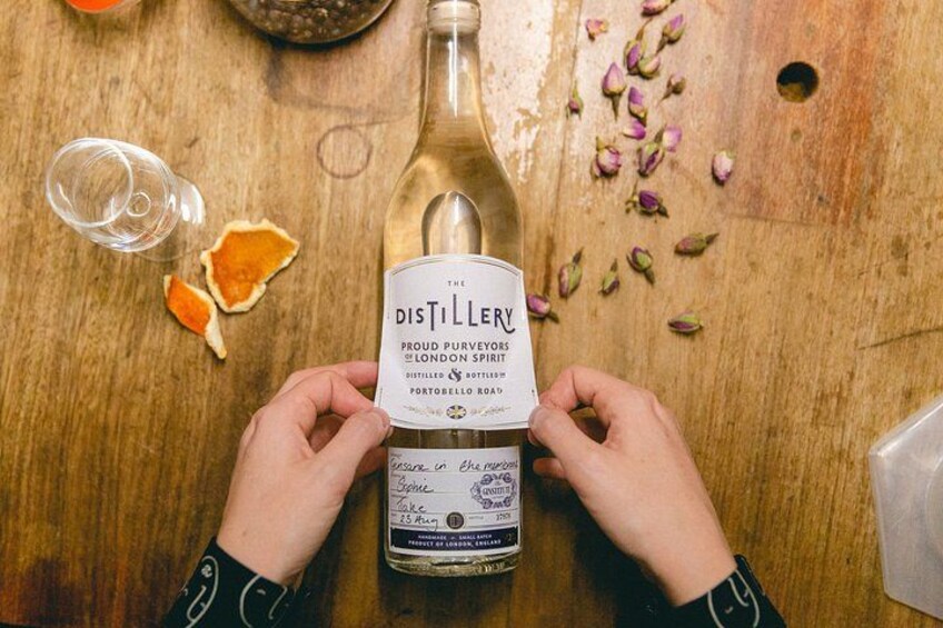 A person holding a bottle of Distillery London spirit on a wooden table with a glass, orange peel, and rose petals. 1