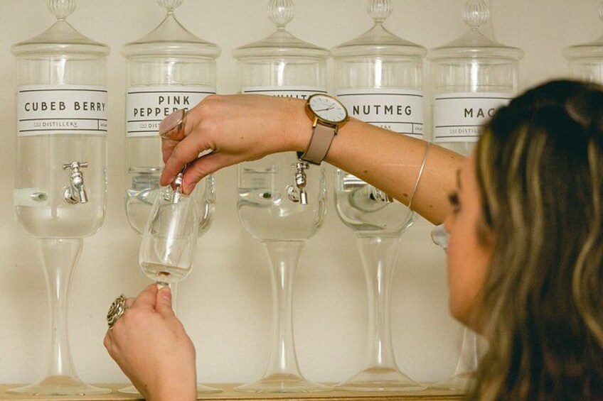 A person pouring liquid from a bottle into a glass. There are jars labeled with different types of spirits like 'CUBEB BERRY' and 'PINK PEPPER.' 4