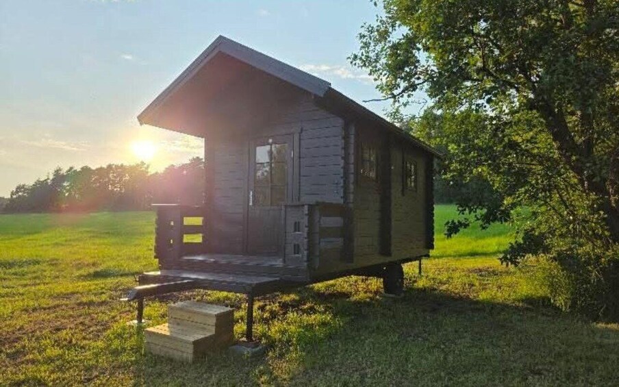 A wooden cabin with a porch, set in a grassy field with trees in the background. 3